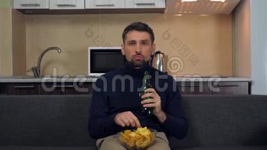 一个穿着深蓝色套衫和灰色裤子的年轻人坐在沙发上，吃薯片，喝啤酒，看电视上的<strong>娱乐节目</strong>